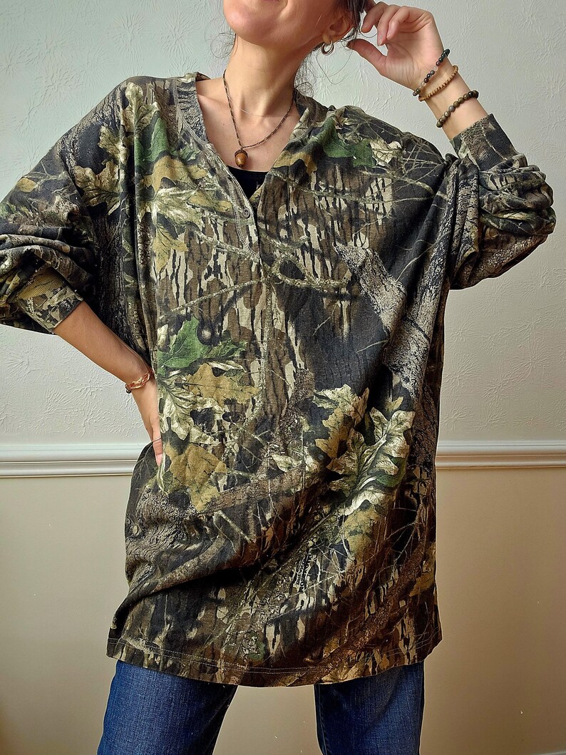 K&ouml;nnte beinhalten: Ein lang&auml;rmeliges Camouflage-Shirt in Braun-, Gr&uuml;n- und Schwarzt&ouml;nen. Das Shirt hat einen V-Ausschnitt und eine lockere Passform. Der Stoff weist ein detailliertes Muster aus Bl&auml;ttern und &Auml;sten auf, das einen Waldeffekt erzeugt. Das Shirt wird mit einer blauen Jeans kombiniert.