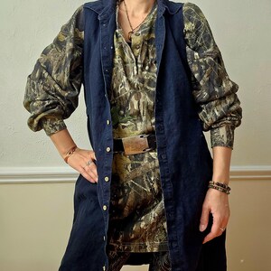 K&ouml;nnte beinhalten: Eine dunkelblaue Jeansweste &uuml;ber einem Hemd und einer Hose mit Camouflage-Muster. Das lang&auml;rmlige Hemd hat eine Knopfleiste und einen Kragen. Die Hose hat ein dekoratives Muster. Ein brauner G&uuml;rtel mit Metallschnalle wird in der Taille getragen.