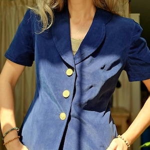 Puede incluir: Una blazer azul marino de manga corta con tres botones dorados. La blazer tiene un cuello clásico y un corte entallado. La modelo lleva un top de color dorado debajo. La parte inferior de la modelo lleva una falda de lunares blancos y negros.