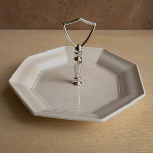 May include: A white, octagonal serving tray with a silver handle. The tray has a crackle glaze finish and a raised geometric design around the edge. The handle is a shield shape.