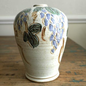 May include: A white ceramic vase with a textured surface and a design of purple grapes and green leaves.