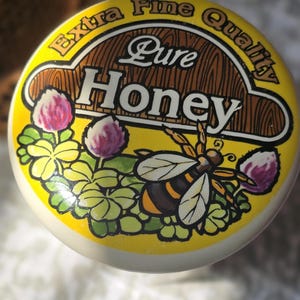 May include: A yellow and brown jar lid with a bee and clover design. The lid says "Extra Fine Quality Pure Honey".