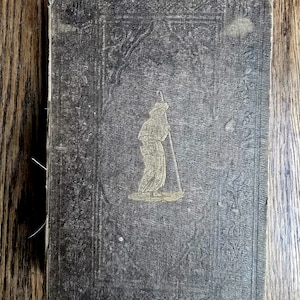 May include: Antique book with a dark gray cover featuring an embossed design of a person with a staff. The cover has a faded appearance with visible wear and tear, suggesting age and use. The book is rectangular and rests on a wooden surface.