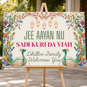 May include: A wedding welcome sign on a wooden easel. The sign has a cream background with floral and peacock designs. Text in green and pink reads "JEE AAYAN NU SADI KURI DA VIAH Dhillon Family Welcomes You."
