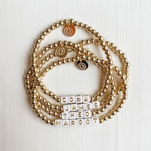 May include: Gold beaded bracelets with white lettered name blocks spelling out CORA, JAMES, THEO, and MARGOT. Each bracelet has a gold sunflower charm. The bracelets are stacked on a white surface.