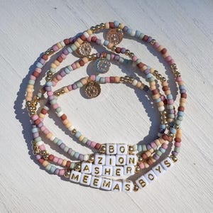 May include: A set of four pastel-colored beaded bracelets. Each bracelet features small, round beads in shades of pink, blue, yellow, and purple. Gold-colored accents and charms with the text "BOYS", "ZION", "ASHER", and "MEEMAS" are also visible.