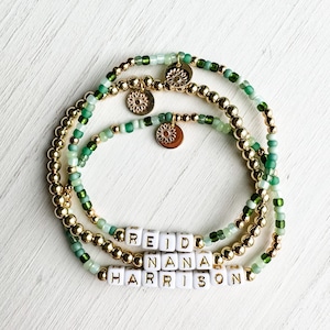 May include: Three beaded bracelets in shades of green and gold. The bracelets feature gold beads, green beads, and small gold charms. The bottom three bracelets spell out the names "REID", "NANA", and "HARRISON" with white letter beads.