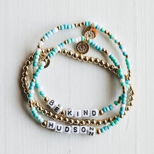 May include: Three beaded bracelets with gold accents and charms. One bracelet spells out "BE KIND" in white letter beads. Another bracelet spells out "HUDSON". The other bracelets have blue, white, and green beads.