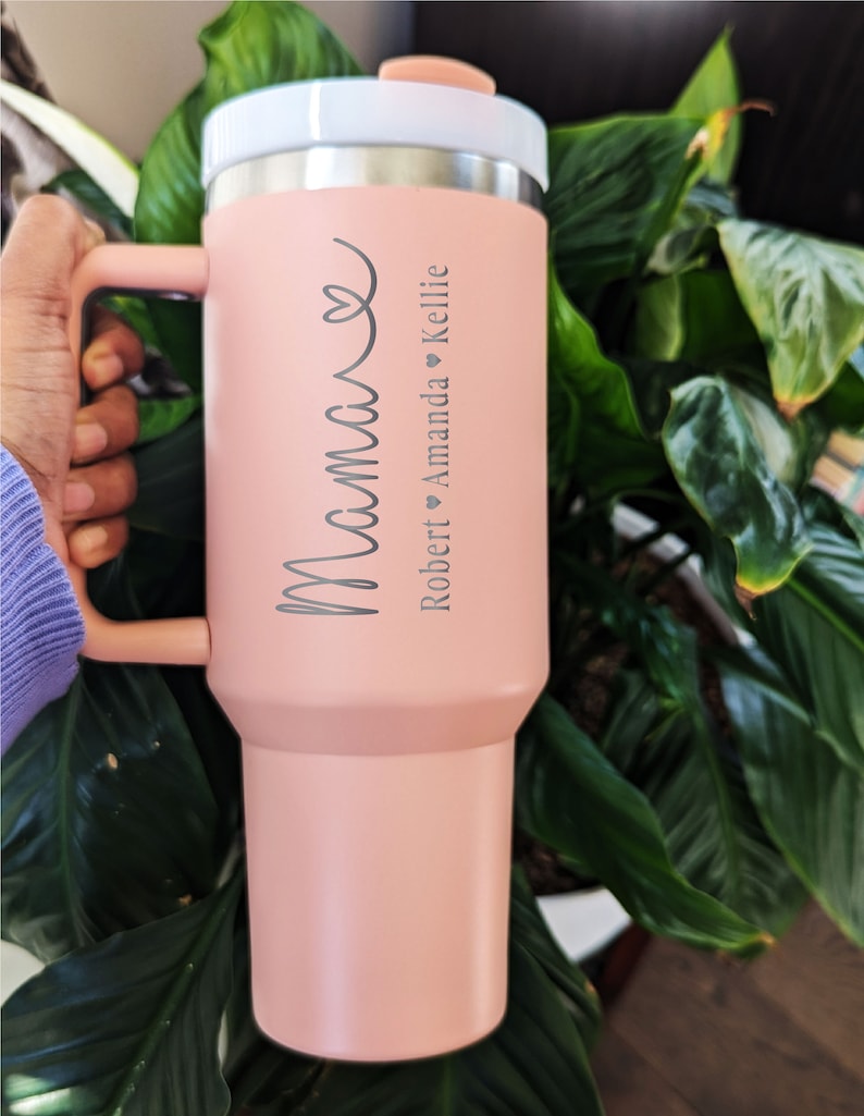 May include: A pink stainless steel tumbler with a handle. The tumbler has the word 'Mama' written on it in a cursive font. Below the word 'Mama' are the names 'Robert', 'Amanda', and 'Kellie' with a heart symbol between each name.