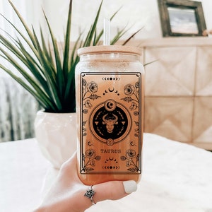 May include: A clear glass bottle with a bamboo lid and a straw. The bottle features a black and white graphic of a bull with the word "TAURUS" printed below it. The graphic is surrounded by a floral design.