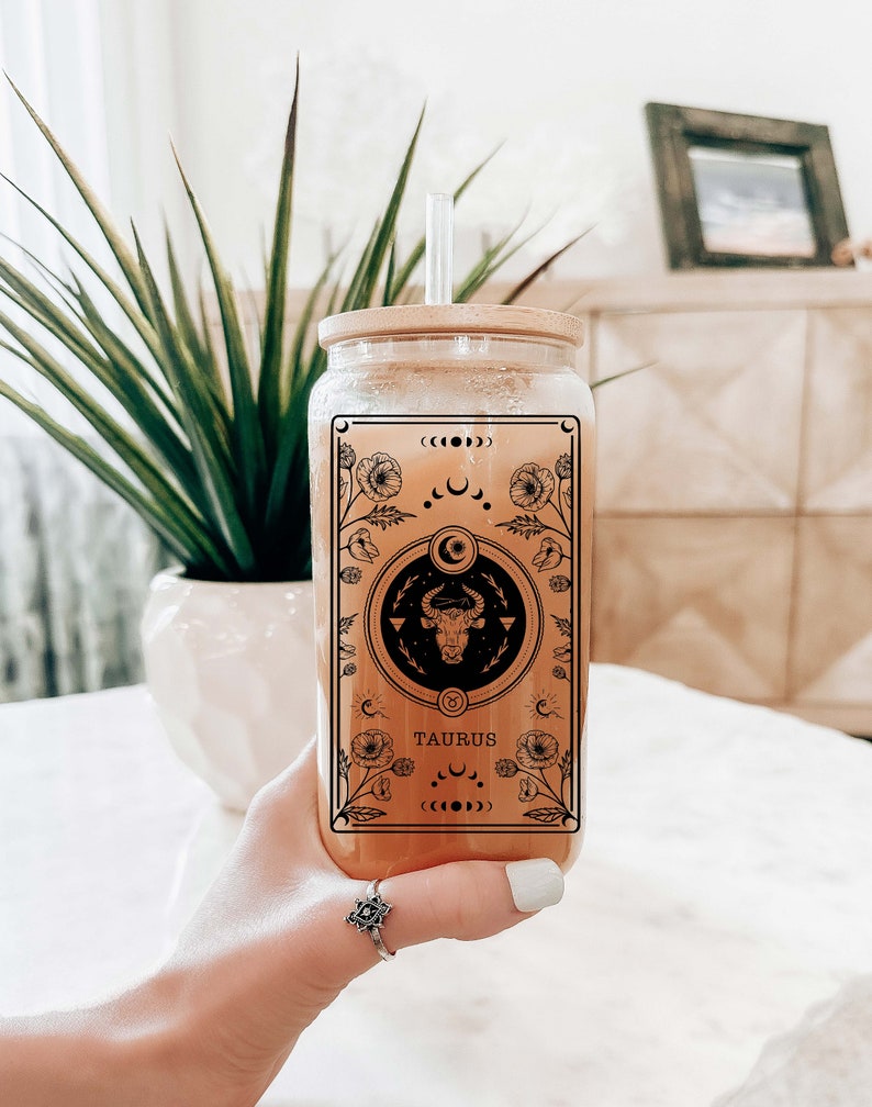 May include: A clear glass bottle with a bamboo lid and a straw. The bottle features a black and white graphic of a bull with the word "TAURUS" printed below it. The graphic is surrounded by a floral design.