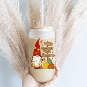 May include: A clear glass tumbler with a wooden lid and a straw. The tumbler has a fall-themed design with a gnome, pumpkins, and a sign that reads "Autumn, Pumpkin Patch, Sweater, Hayride".