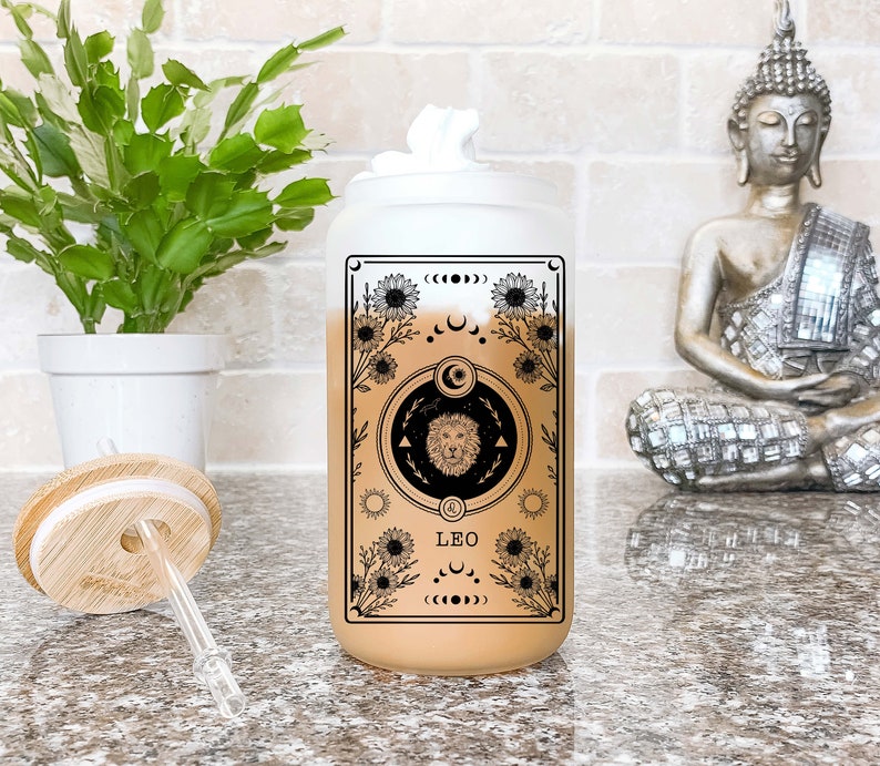 May include: A clear glass tumbler with a black and white design featuring a lion, flowers, and the word "Leo". The tumbler has a bamboo lid and a straw.