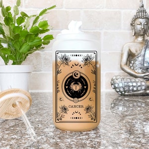 May include: A frosted glass tumbler with a black and white design featuring the astrological sign Cancer. The design includes a crab, flowers, and moons. The tumbler has a bamboo lid and a clear straw.