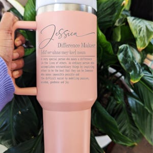 May include: A pink stainless steel travel mug with a white lid and a black handle. The mug has the name "Jessica" and the text "Difference Maker [dif-er-uhns-mey-ker] noun" engraved on it. The text describes a difference maker as a special person who inspires others to be their best.
