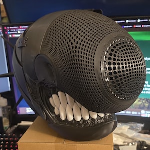 May include: A black helmet featuring a textured, mesh-like front panel with a rounded, protruding snout. The lower section displays a row of prominent, white, tooth-like structures set against a dark, glossy base, creating a distinct, creature-inspired appearance.