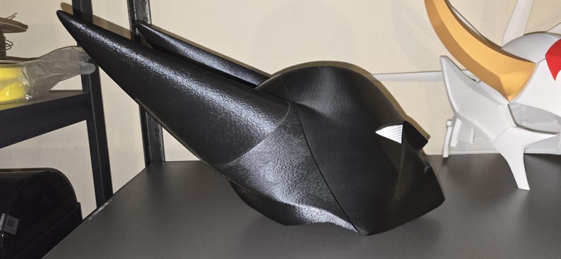 K&ouml;nnte beinhalten: Ein schwarzer Helm mit zwei langen, spitzen, nach oben gerichteten Ohren und einer glatten, konturierten Oberfl&auml;che. Der Helm hat ein markantes, kantiges Design mit einer hervorstehenden Nase und einer kleinen, wei&szlig;en, dreieckigen Augen&ouml;ffnung.