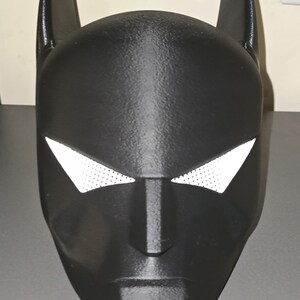 K&ouml;nnte beinhalten: Eine schwarze Vollkopfmaske mit zwei hohen, spitzen Ohren an der Oberseite und scharfen, kantigen Augenausschnitten. Die Augen&ouml;ffnungen sind mit einem wei&szlig;en, feinmaschigen Material bedeckt. Die Maske hat eine matte Oberfl&auml;che und ein spitzes Kinn.
