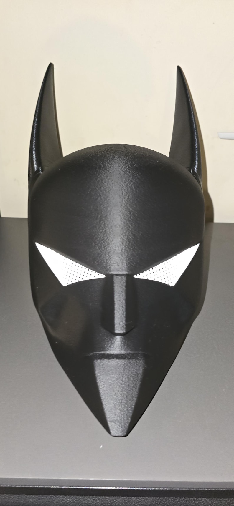 K&ouml;nnte beinhalten: Eine schwarze Vollkopfmaske mit zwei hohen, spitzen Ohren an der Oberseite und scharfen, kantigen Augenausschnitten. Die Augen&ouml;ffnungen sind mit einem wei&szlig;en, feinmaschigen Material bedeckt. Die Maske hat eine matte Oberfl&auml;che und ein spitzes Kinn.