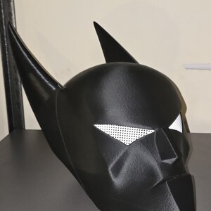 K&ouml;nnte beinhalten: Eine mattschwarze Superheldenmaske mit zwei hohen, spitzen Ohren und wei&szlig;en Netz-Augenabdeckungen. Die Maske hat ein strukturiertes, kantiges Design mit einem definierten Nasensteg und einem spitzen Kinnbereich sowie einer 3D-Druck-Textur.