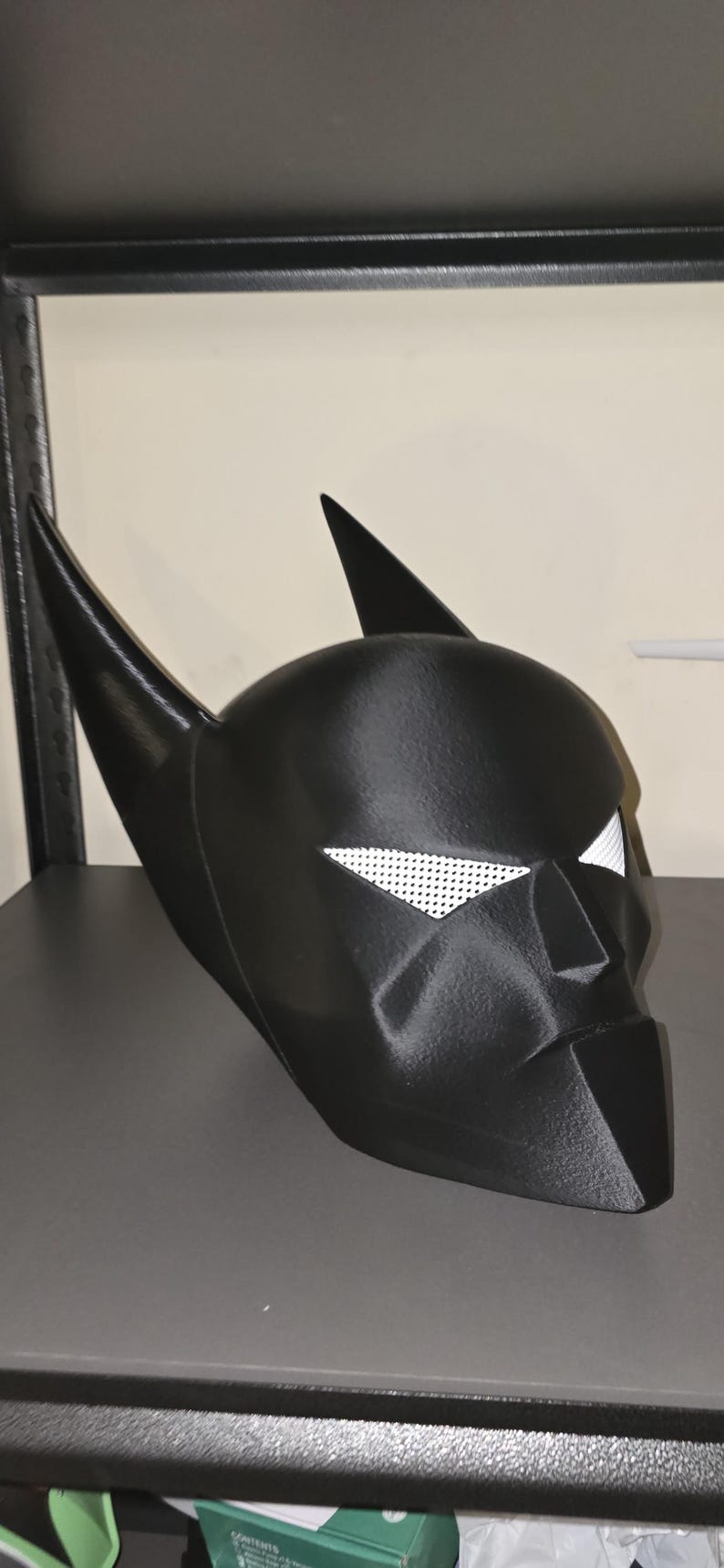 K&ouml;nnte beinhalten: Eine mattschwarze Superheldenmaske mit zwei hohen, spitzen Ohren und wei&szlig;en Netz-Augenabdeckungen. Die Maske hat ein strukturiertes, kantiges Design mit einem definierten Nasensteg und einem spitzen Kinnbereich sowie einer 3D-Druck-Textur.