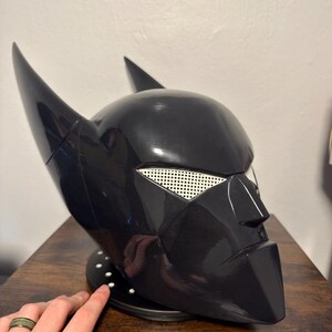 K&ouml;nnte beinhalten: Ein gl&auml;nzender schwarzer Vollhelm mit zwei markanten spitzen Ohren, einem scharfen, kantigen Nasensteg und wei&szlig;en Netzabdeckungen f&uuml;r die Augen. Der Helm ist auf einem runden schwarzen Sockel mit kleinen wei&szlig;en kreisf&ouml;rmigen Akzenten montiert.