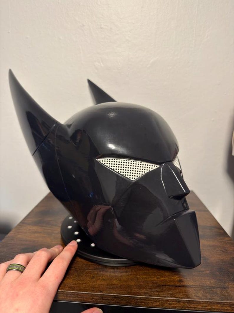 K&ouml;nnte beinhalten: Ein gl&auml;nzender schwarzer Vollhelm mit zwei markanten spitzen Ohren, einem scharfen, kantigen Nasensteg und wei&szlig;en Netzabdeckungen f&uuml;r die Augen. Der Helm ist auf einem runden schwarzen Sockel mit kleinen wei&szlig;en kreisf&ouml;rmigen Akzenten montiert.