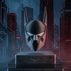 K&ouml;nnte beinhalten: Ein schwarz-wei&szlig;er Batman Beyond Helm mit spitzen Ohren und einem schlanken, kantigen Design. Der Helm ist auf einem rechteckigen Sockel mit einem Fledermaus-Emblem ausgestellt, wobei der Text BATMAN BEYOND HELMET unten aufgedruckt ist.
