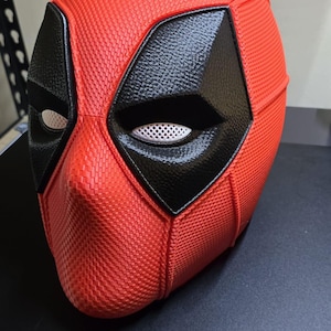 May include: A red and black Deadpool mask. The mask is predominantly red with a textured pattern, and features black eye and mouth details. The eye openings have white mesh. The mask is a replica of the character's mask from the Marvel comics and films.