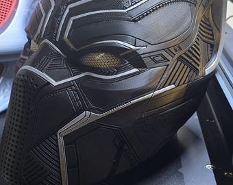 Könnte beinhalten: Eine detaillierte schwarze und silberne Black Panther-Maske mit komplizierten geometrischen Mustern. Die Maske hat Augen- und Mundöffnungen und ist wahrscheinlich ein Cosplay- oder Sammlerobjekt. Die Maske liegt auf einer schwarzen Oberfläche.