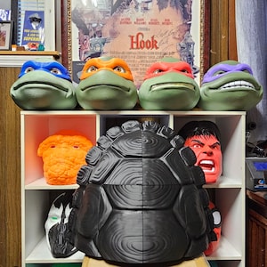 May include: A collection of four Teenage Mutant Ninja Turtles character heads in blue, orange, red, and purple masks, displayed above a black turtle shell, with additional character masks including an orange rock-like figure and a red-faced figure.