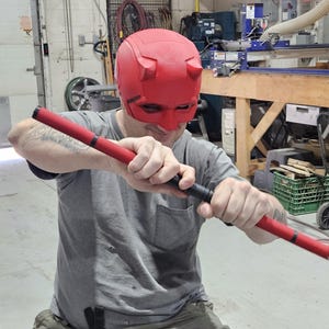 May include: A red superhero mask with horns, paired with a red and black baton. The mask covers the eyes and forehead. The baton is held in both hands. The person is wearing a gray t-shirt and khaki pants.