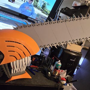 May include: A 3D printed chainsaw head prop featuring a bright orange casing, a long grey blade with sharp teeth along the edge, and a set of white pointed teeth protruding from the lower section. The design includes a grey handle and vents.