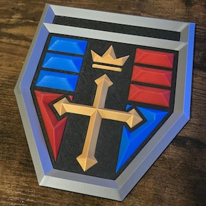 May include: A shield-shaped emblem with a silver border. The design features a gold cross, a gold crown, and blue and red geometric shapes on a black background. The emblem has a 3D appearance.