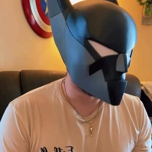 K&ouml;nnte beinhalten: Eine dunkelgraue, kantige Vollkopfmaske mit hohen, spitzen Ohren und einem geometrischen Gesicht. Die Maske hat eine glatte, matte Oberfl&auml;che mit deutlichen Paneellinien und wei&szlig;en Augenausschnitten. Die Person tr&auml;gt ein beiges T-Shirt mit dem aufgedruckten Text "Football DAD" auf der Brust, zusammen mit einer kleinen Football-Grafik und einer goldfarbenen Kette.