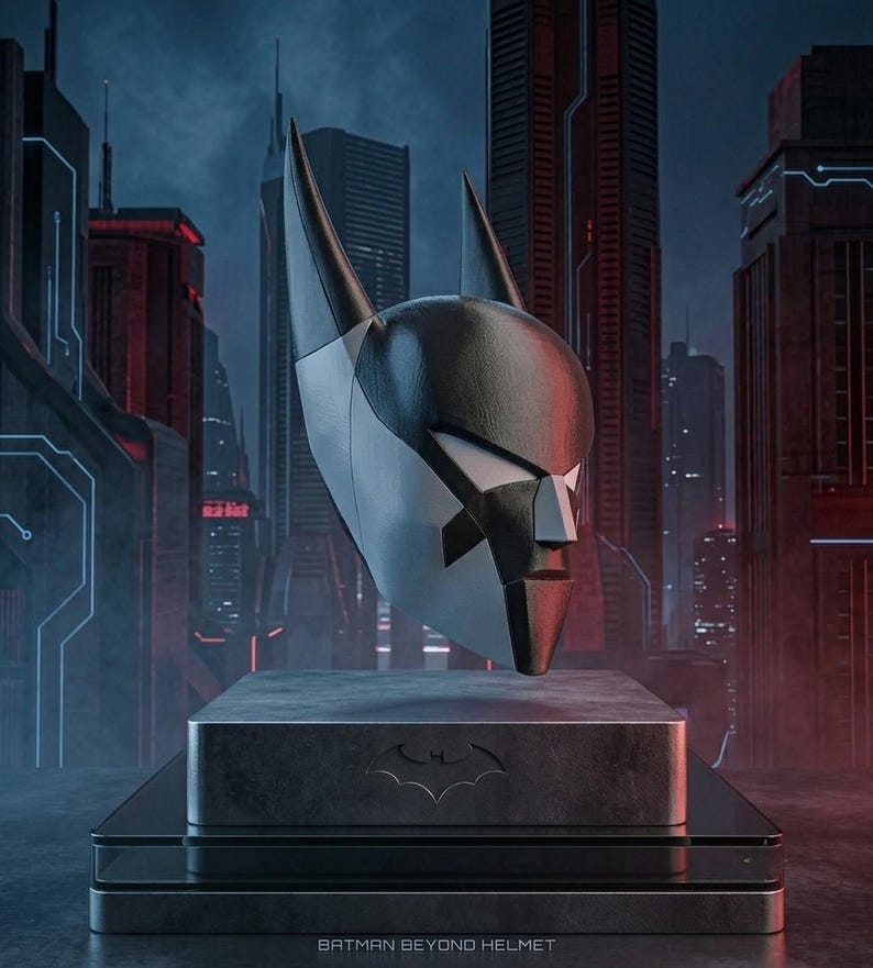K&ouml;nnte beinhalten: Ein schwarz-silberner Batman Beyond Helm mit spitzen Ohren und einer stilisierten Gesichtsmaske, montiert auf einem rechteckigen Sockel mit Fledermaus-Emblem. Der Sockel tr&auml;gt unten mittig den Schriftzug BATMAN BEYOND HELMET.