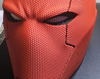May include: A full-head helmet featuring a vibrant red exterior with a distinct, fine-textured, dotted surface pattern. The helmet includes narrow, angular eye openings with white mesh inserts and a sharp, defined jawline with a horizontal seam.