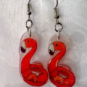 May include: A pair of earrings featuring red flamingo pool float designs. The earrings are made of clear plastic with silver hooks.