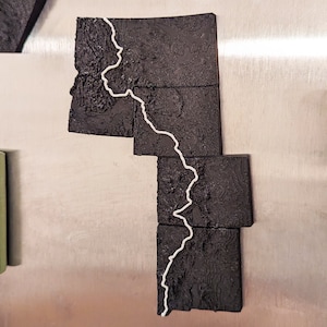 May include: A black and white map of a state, possibly Maine, with a white line tracing a path through the state. The map is made of individual pieces that fit together.