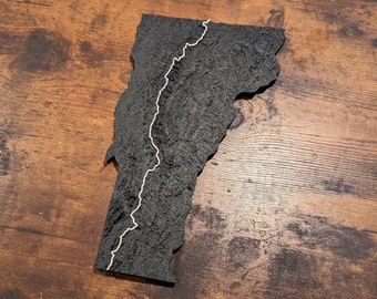 May include: A black, textured, 3D map of Vermont with a white line tracing the Long Trail hiking route.