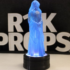 May include: A blue translucent 3D printed figurine of a person wearing a robe, standing on a black base with a light underneath. The figurine is lit from below, casting a blue glow. The background is black with the text "RK Props" in white.
