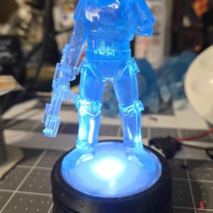 May include: A blue, translucent Stormtrooper figurine, lit from below, stands on a black base. The figure is holding a blaster rifle and has a detailed helmet. The base emits a bright, white light, creating a glowing effect.