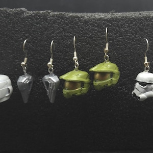 May include: A selection of handmade earrings showcasing miniature helmet designs from popular culture. The earrings include white Stormtrooper helmets, green Halo Master Chief helmets, and other sci-fi inspired designs. Each earring is suspended from a silver-coloured hook.