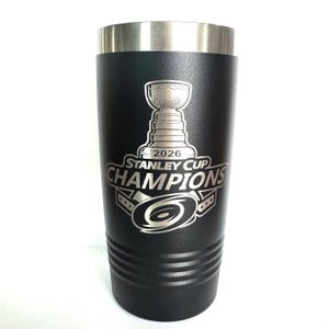 K&ouml;nnte beinhalten: Ein mattschwarzer Edelstahlbecher mit silbernem Rand. Die Vorderseite zeigt eine Gravur des Stanley Cup Pokals &uuml;ber dem Text 2026 STANLEY CUP CHAMPIONS und ein kreisf&ouml;rmiges Hurrikan-Logo mit drei Sternen auf jeder Seite.