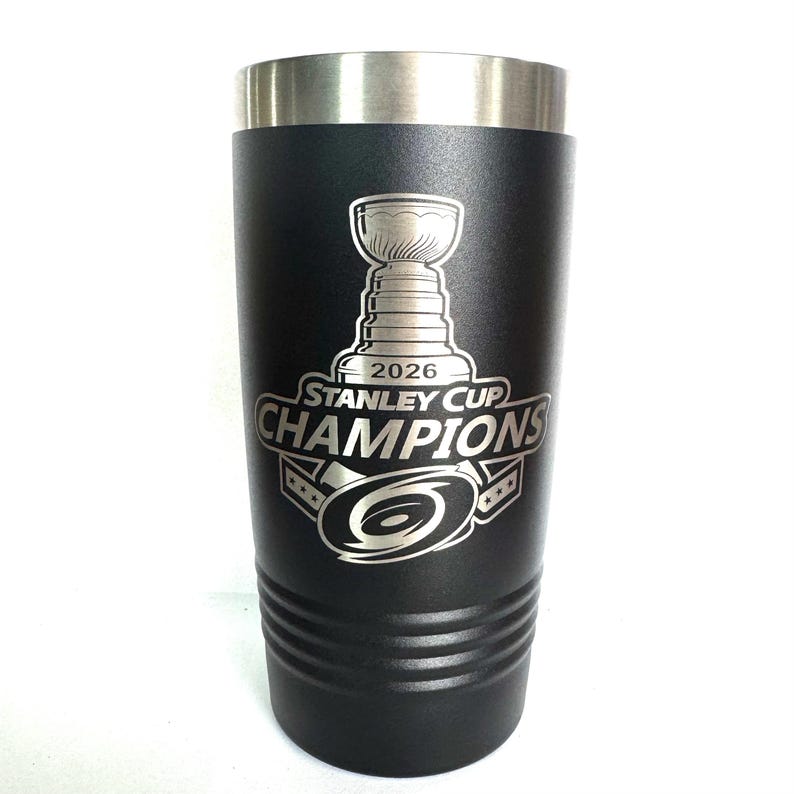 K&ouml;nnte beinhalten: Ein mattschwarzer Edelstahlbecher mit silbernem Rand. Die Vorderseite zeigt eine Gravur des Stanley Cup Pokals &uuml;ber dem Text 2026 STANLEY CUP CHAMPIONS und ein kreisf&ouml;rmiges Hurrikan-Logo mit drei Sternen auf jeder Seite.