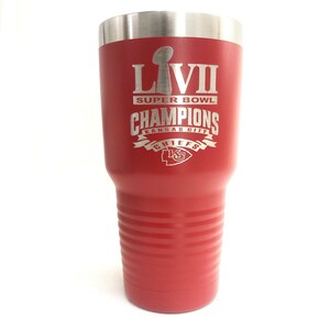 May include: A red insulated tumbler with a silver rim. The tumbler features the text "LVII SUPER BOWL CHAMPIONS KANSAS CITY CHIEFS" in silver lettering, along with the team's logo. The tumbler has a ridged design.