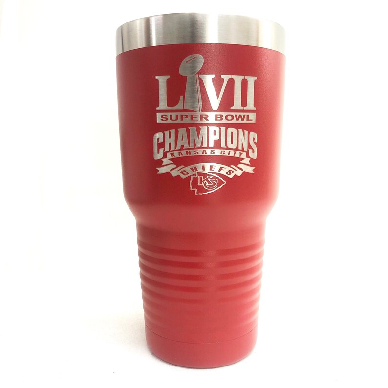 May include: A red insulated tumbler with a silver rim. The tumbler features the text "LVII SUPER BOWL CHAMPIONS KANSAS CITY CHIEFS" in silver lettering, along with the team's logo. The tumbler has a ridged design.