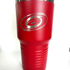 K&ouml;nnte beinhalten: Ein leuchtend roter, pulverbeschichteter Edelstahlbecher mit einer texturierten Oberfl&auml;che und einem silberfarbenen, lasergravierten Carolina Hurricanes-Logo auf der Vorderseite. Der Becher hat einen glatten Metallrand und horizontale Rillen am Boden.