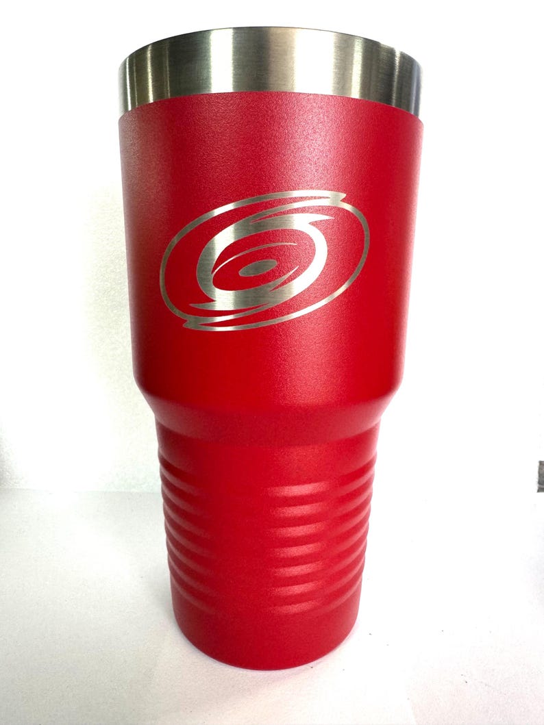 K&ouml;nnte beinhalten: Ein leuchtend roter, pulverbeschichteter Edelstahlbecher mit einer texturierten Oberfl&auml;che und einem silberfarbenen, lasergravierten Carolina Hurricanes-Logo auf der Vorderseite. Der Becher hat einen glatten Metallrand und horizontale Rillen am Boden.