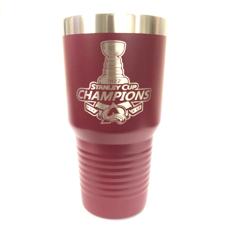 May include: A maroon stainless steel tumbler with a silver lid. The tumbler has an engraved design of the Stanley Cup, the text "2022 Stanley Cup Champions", and the Colorado Avalanche logo.